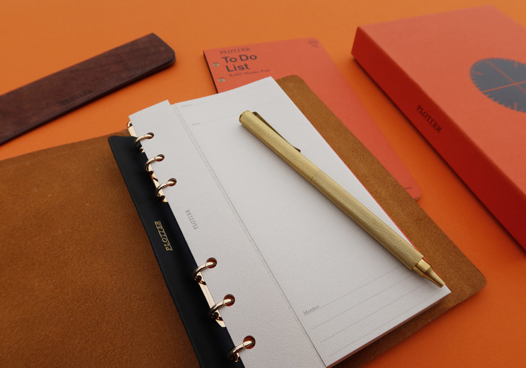 A PLOTTER Leather Binder opens to a blank page with different stationery accessories around it.