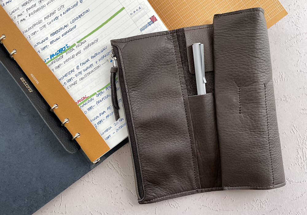 A Roll Pen Case with a PLOTTER pen in one of its slot, half folded over on top of an opened PLOTTER. 