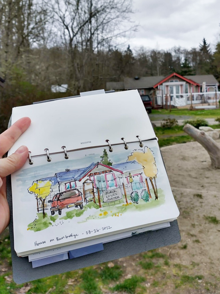A hand holding an open PLOTTER with a drawing of a house on Bainbridge.