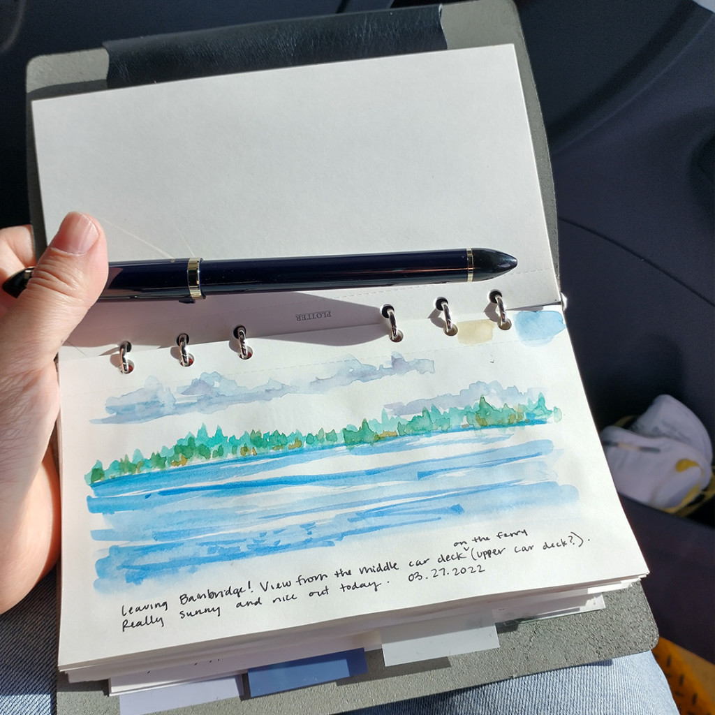 A hand holding open a PLOTTER with a painting of a water scene from the ferry.