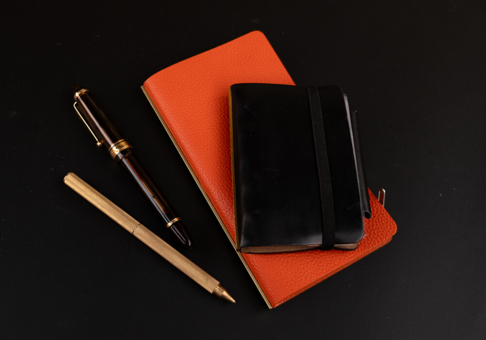 A display of PLOTTER Leather Binder in Orange Shrink, Black Liscio, with a fountain pen and a PLOTTER ballpoint pen.