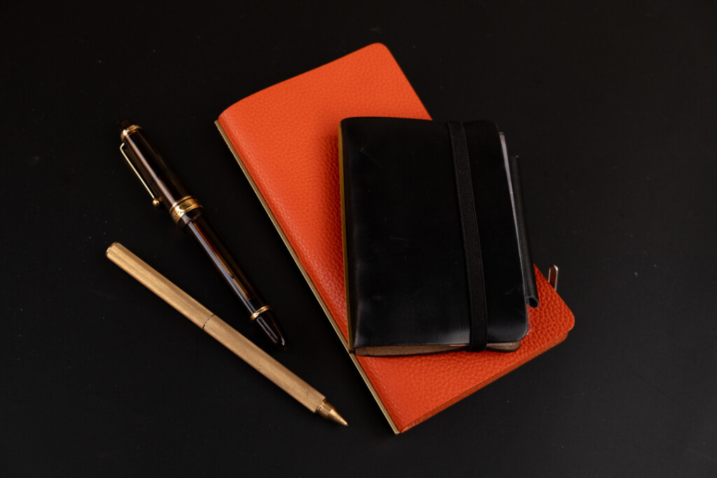 A display of PLOTTER Leather Binder in Orange Shrink, Black Liscio, with a fountain pen and a PLOTTER ballpoint pen.