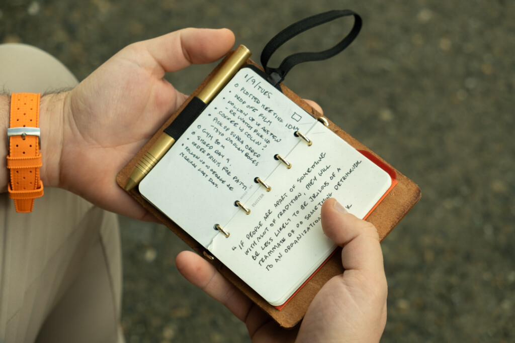 Chris Giannakos holding his open PLOTTER Mini 5 leather binder to a notes page with task lists and bullet points.