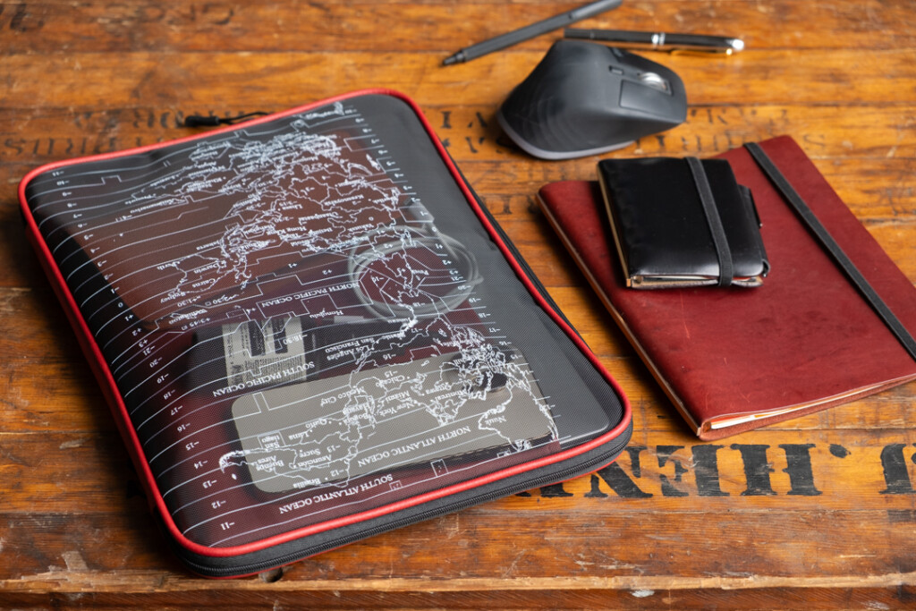 A red PLOTTER A4 Mesh Zipper Case is being used as a laptop case beside other tech devices and stationery.