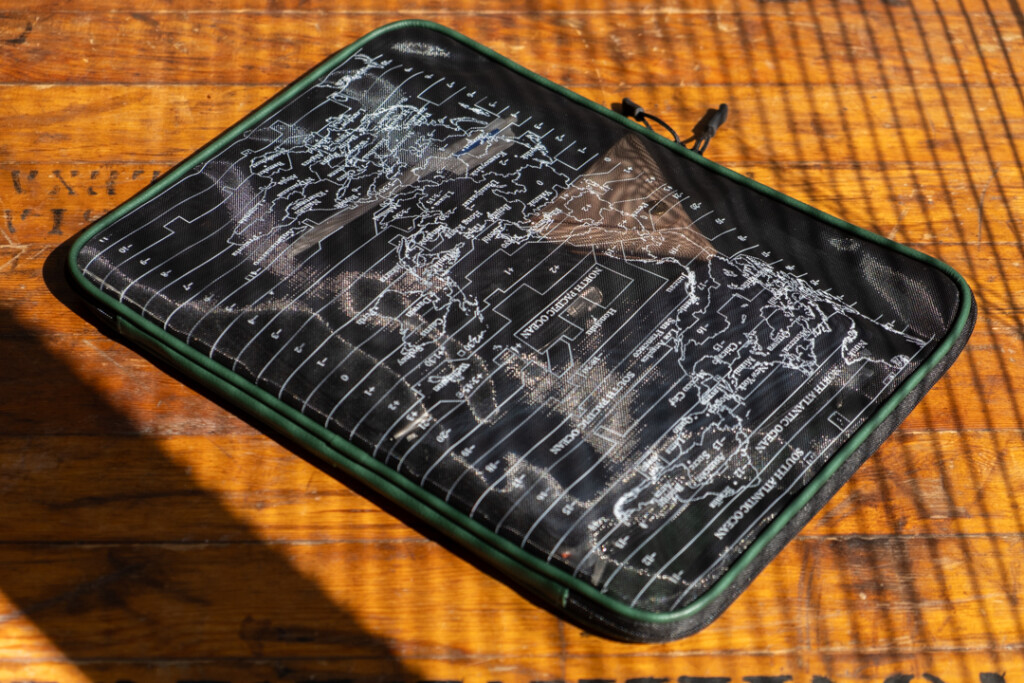 A green PLOTTER A4 Mesh Zipper Case is being used as a laptop case beside other tech devices and stationery.