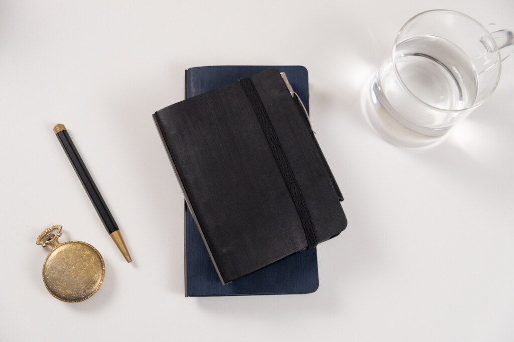 A PLOTTER Horse Hair II Narrow and Mini Size Leather Binder on top of each other on a white table with a cup of water and writing utensils beside it.