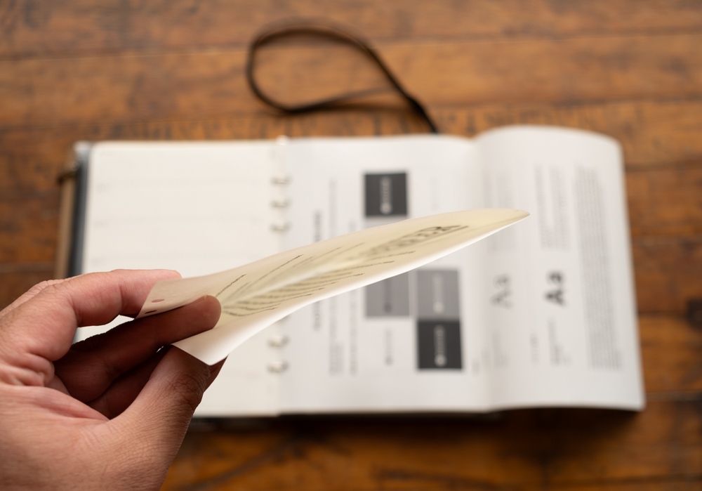 Hand holding US-Letter size paper folded in half in front of a PLOTTER leather binder.