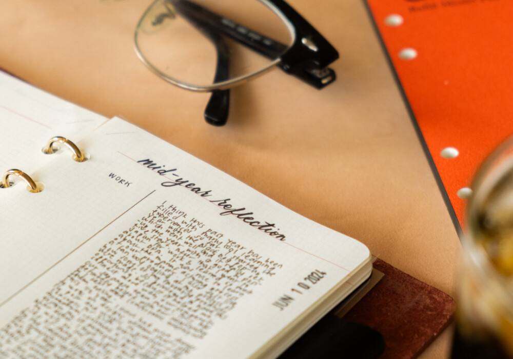 Close-up of PLOTTER Leather Binder open with notes beside ice coffee, Refill Memo Pad, Pen and eye glasses.