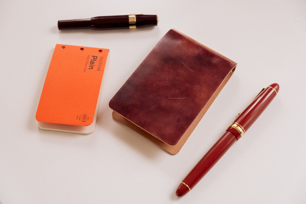 PLOTTER 3-Ring Leather Binder closed beside a refill memo pad, red fountain pen, and pocket pen.