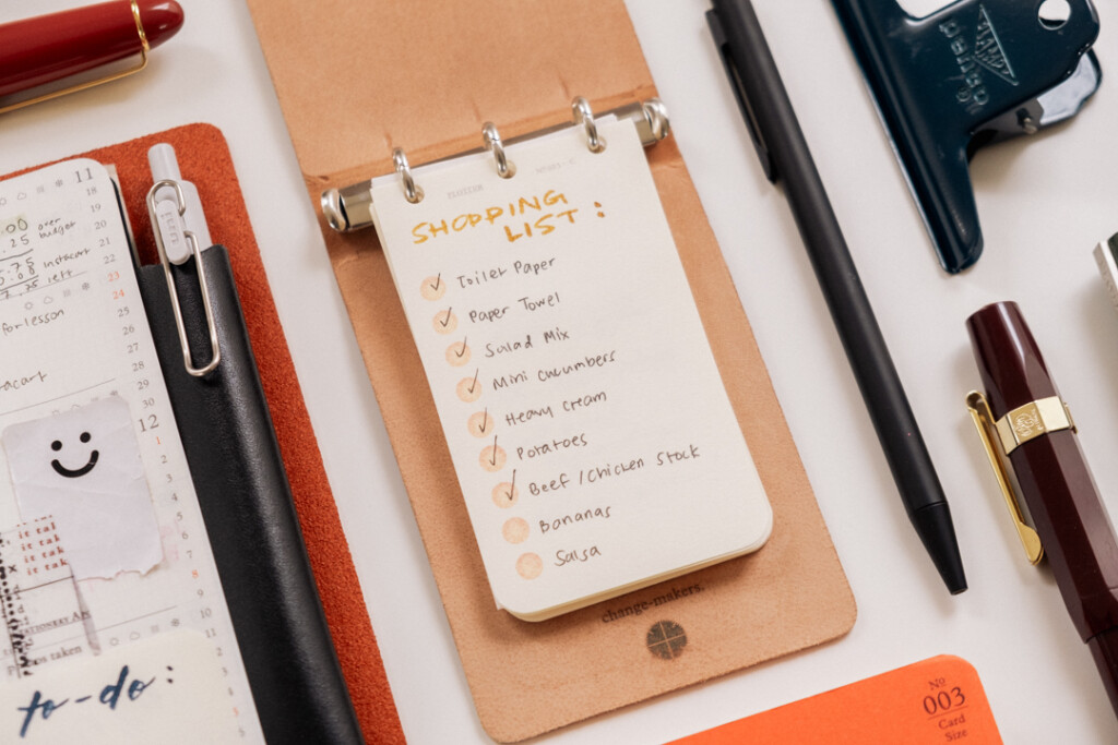 PLOTTER 3-Ring Leather Binder open to a shopping list of groceries beside other stationery.