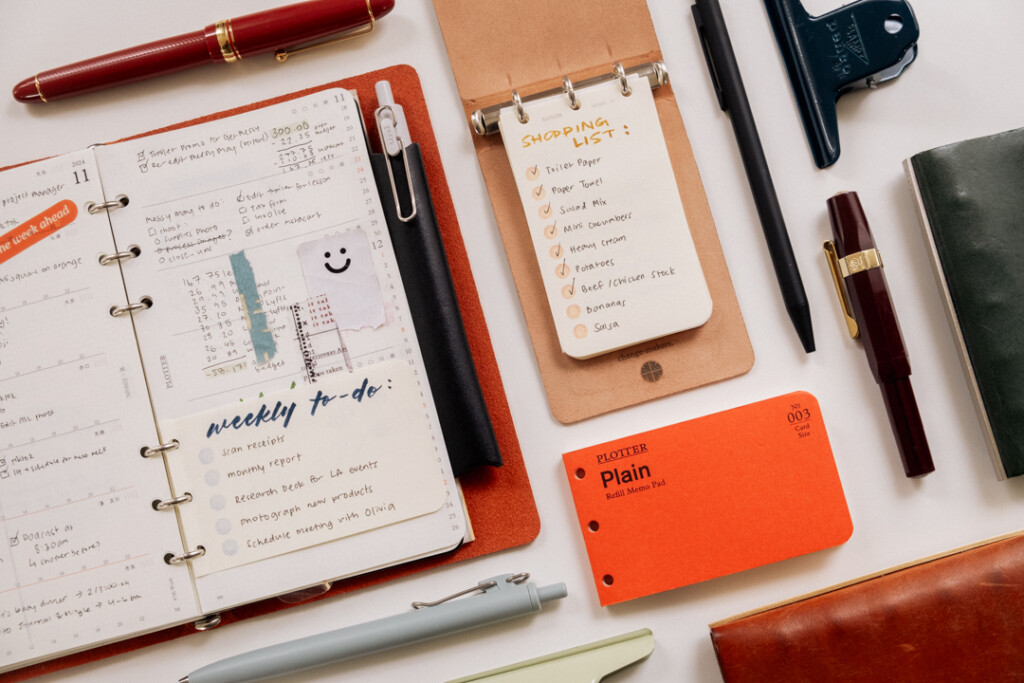 PLOTTER 3-Ring Leather Binder open to a shopping list of groceries beside other stationery and a PLOTTER Bible Size Leather Binder.