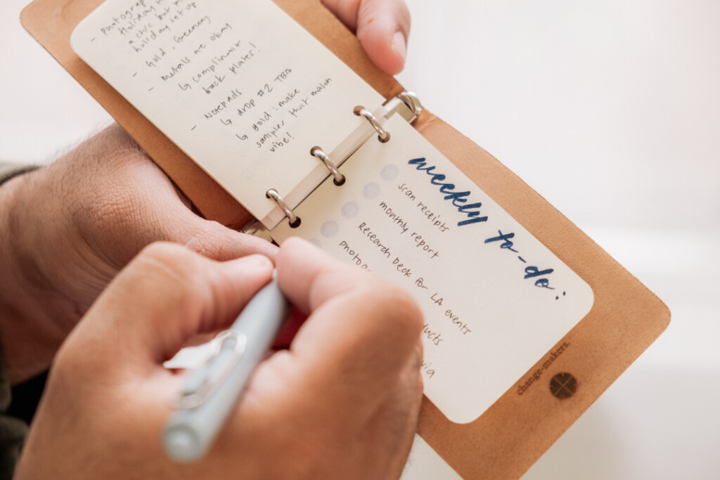 A person writing a weekly to-do list in a PLOTTER 3-Ring Leather Binder with a gel pen.
