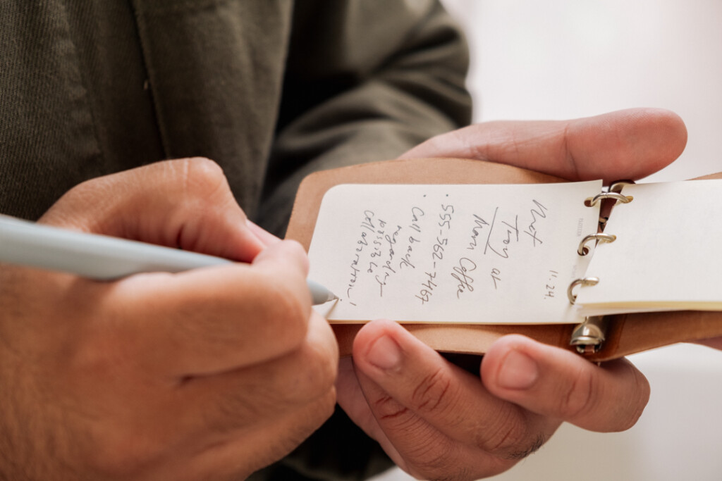 A person writing notes in a PLOTTER 3-Ring Leather Binder with a gel pen.