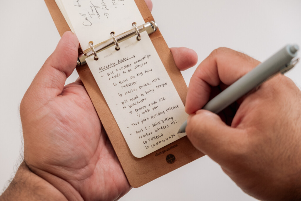 A person writing meeting notes in a PLOTTER 3-Ring Leathe Binder with a gel pen.
