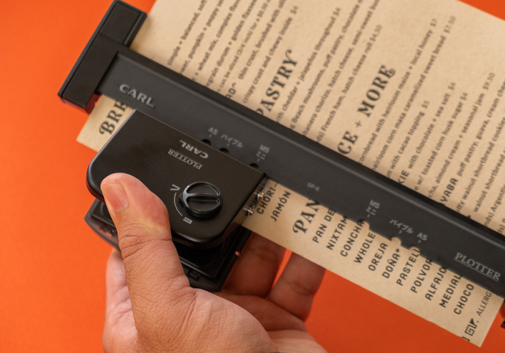 A hand holding the PLOTTER x CARL Refill Maker Hole Punch and punching holes in a menu flyer.