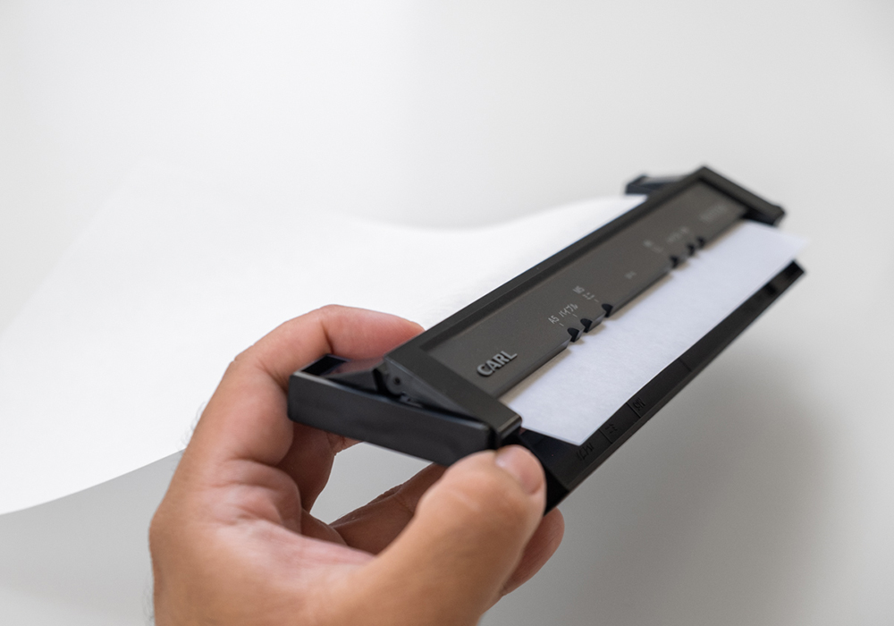 A hand holding the gauge section of the Refill Maker Hole Punch and the paper sliding through the feed.