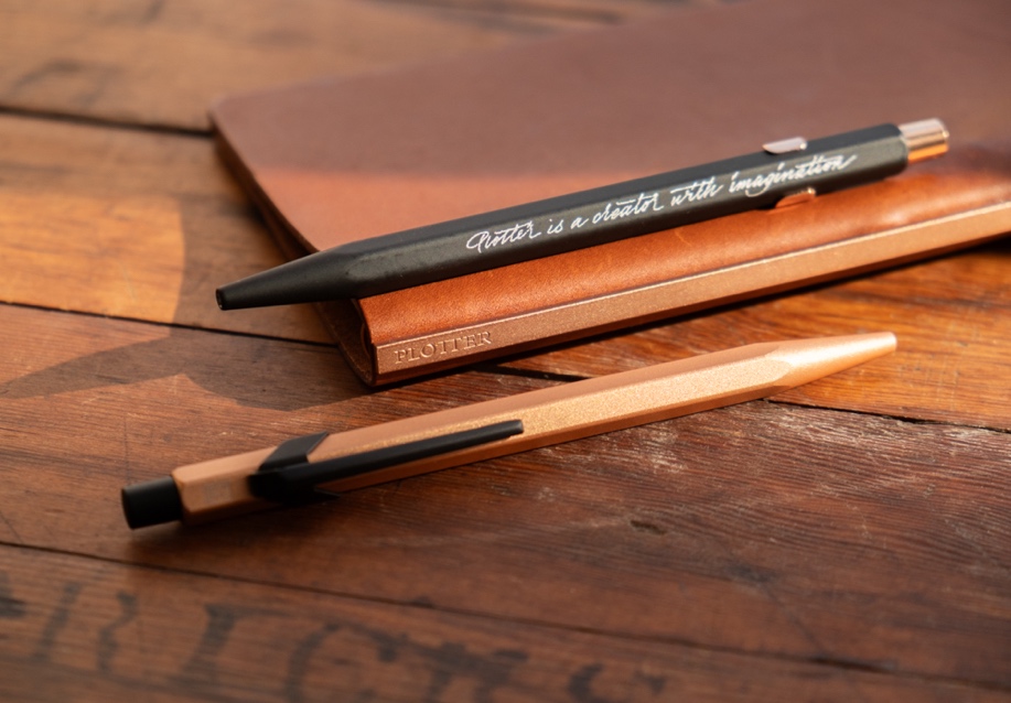The PLOTTER x CARAN D’ACHE 849 Ballpoint Pen in black and rose gold on a wooden table.