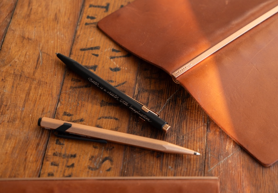 The PLOTTER x CARAN D’ACHE 849 Ballpoint Pen in black and rose gold on a wooden table.