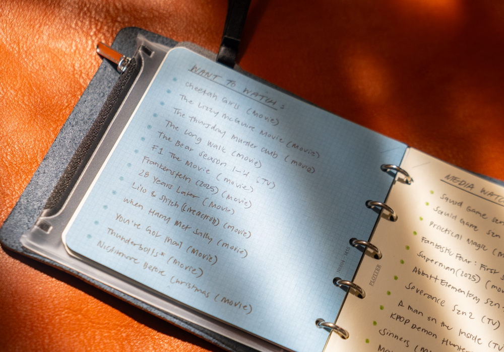 TV and Movie notes and watchlist inside a PLOTTER Mini 5 Square Size Leather Binder on an orange leather background in high-contrast lighting.