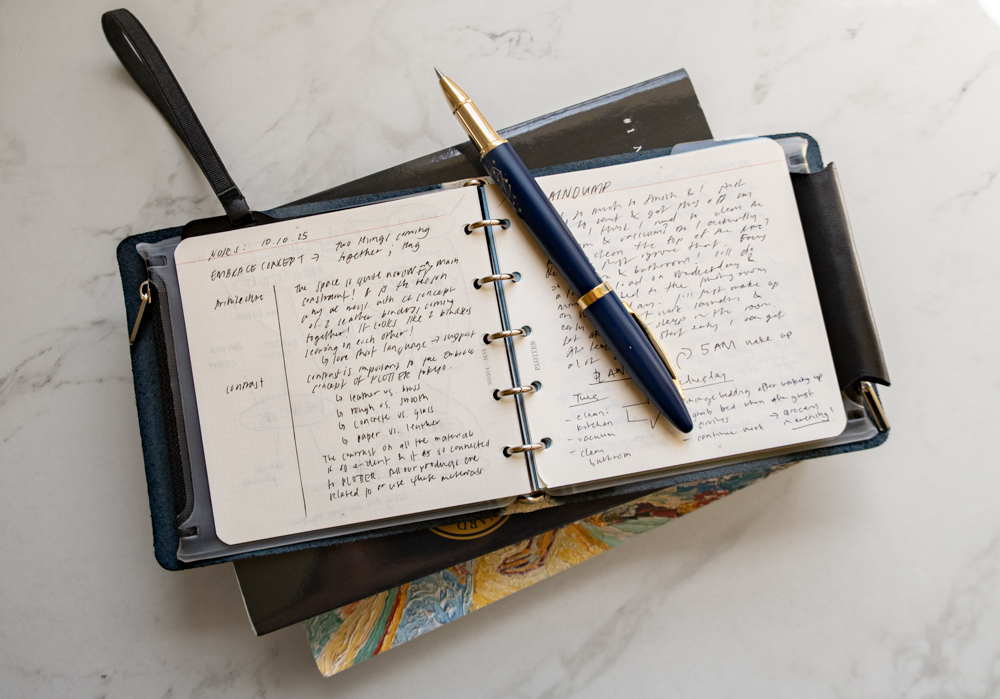 Stream of consciousness notes and journal reflections inside a PLOTTER Mini 5 Square Size Leather Binder beside a pen and books.