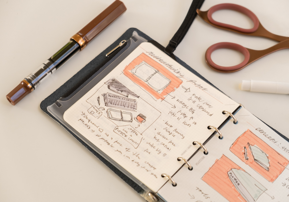 Sketches inside a PLOTTER Mini 5 Square Leather Binder about photography and shooting notes, beside a scatter of pens and writing tools.