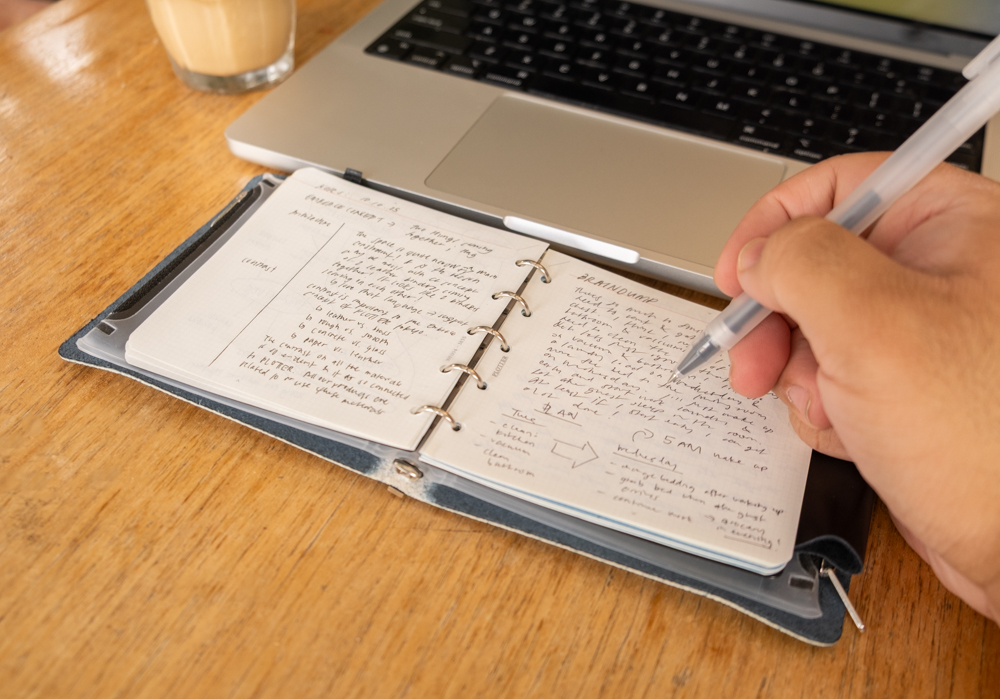 A person writing in a PLOTTER Mini 5 Square leather binder beside a laptop and iced coffee.