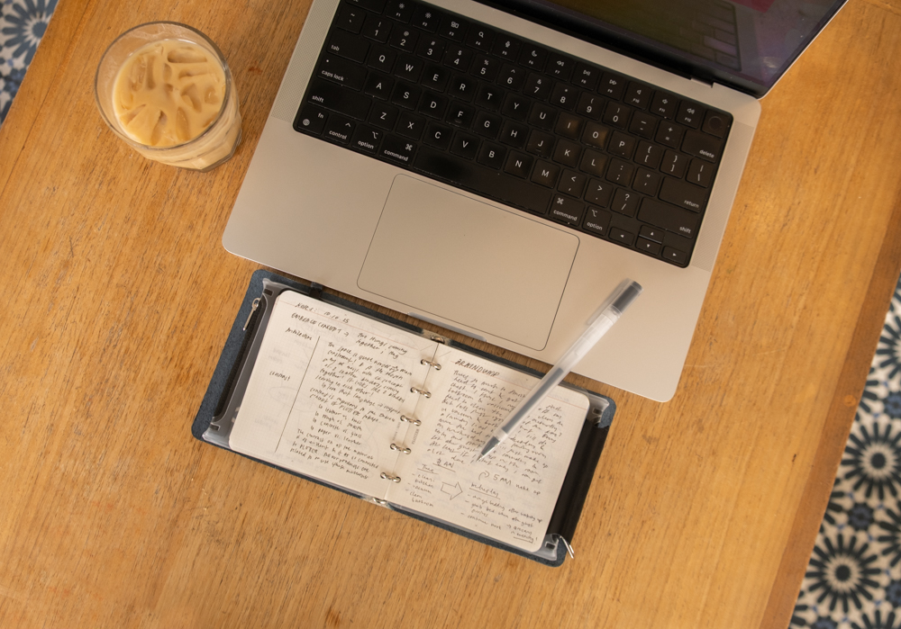 A PLOTTER Mini 5 Square Leather Binder propped up against a laptop with notes filled in the binder; there is an iced coffee and pen beside the laptop and leather binder.