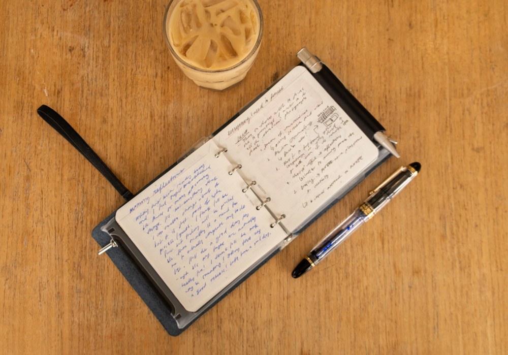 Overhead shot of a person's journal reflections inside a PLOTTER Mini 5 Square Size Leather Binder beside a fountain pen and iced coffee on a wooden table.