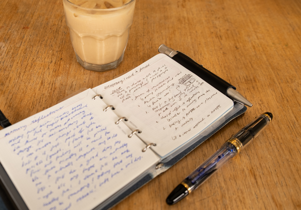 Journal reflections inside a PLOTTER Mini 5 Square Size Leather Binder beside a fountain pen and iced coffee on a wooden table.