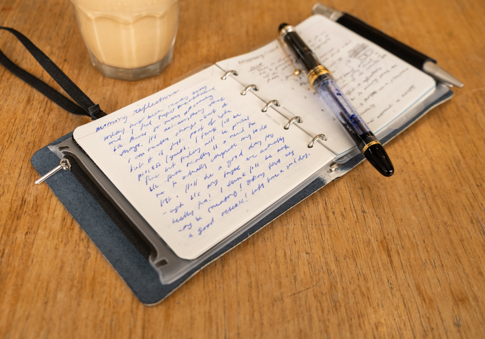 Journal reflections inside a PLOTTER Mini 5 Square Size Leather Binder beside a fountain pen and iced coffee on a wooden table.