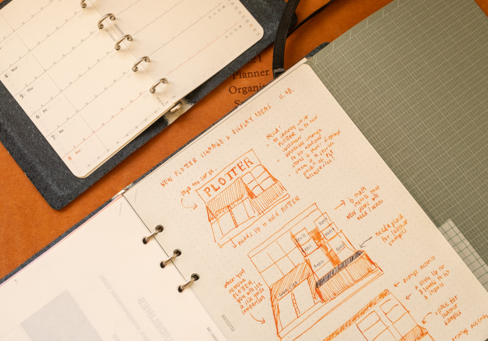 A PLOTTER Leather Binder open to a page with sketches and diagrams for a shop display plan. The sketches are made in orange ink on the PLOTTER 5 mm Dot Grid Refill paper.