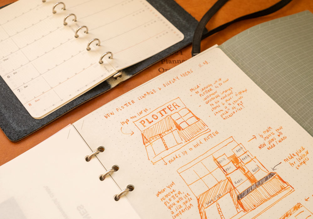 A PLOTTER Leather Binder open to a page with sketches and diagrams for a shop display plan. The sketches are made in orange ink on the PLOTTER 5 mm Dot Grid Refill paper.