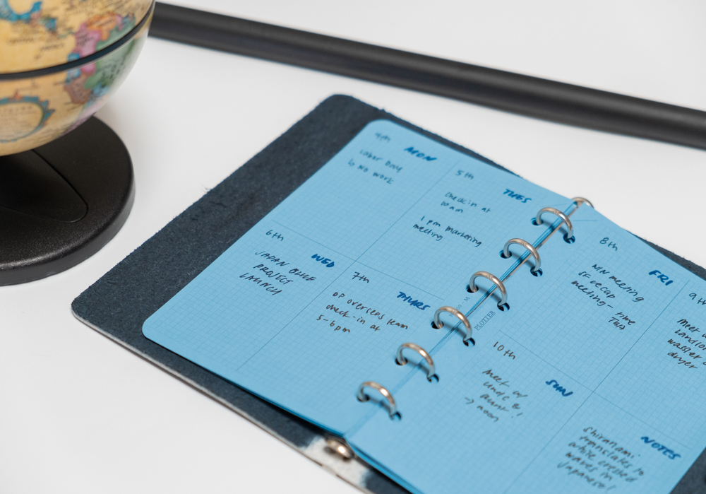 Open blue planner with handwritten notes, showing a weekly schedule. A globe is partially visible on the left, suggesting a workspace context.