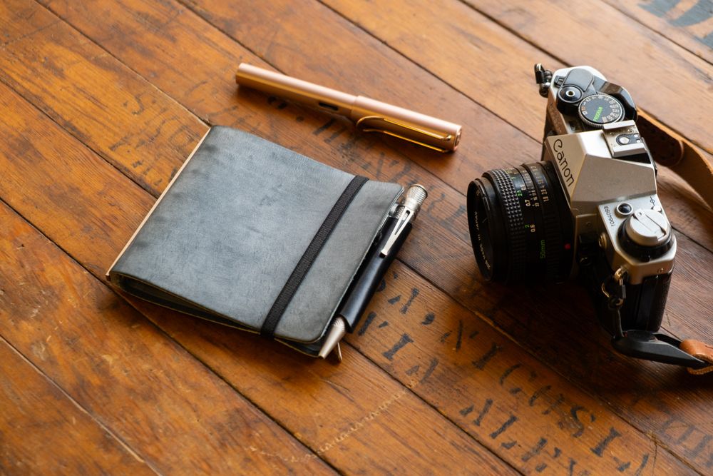 PLOTTER Shiranami Leather Binder in Mini 5 Square next to a fountain pen and a camera.