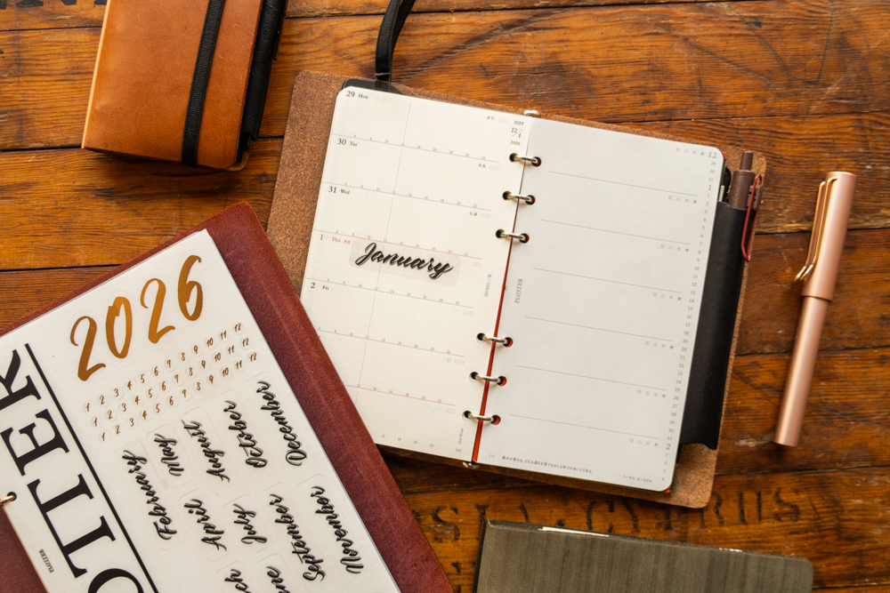 An opened PLOTTER leather binder next to a sticker sheet with the theme of 2026.