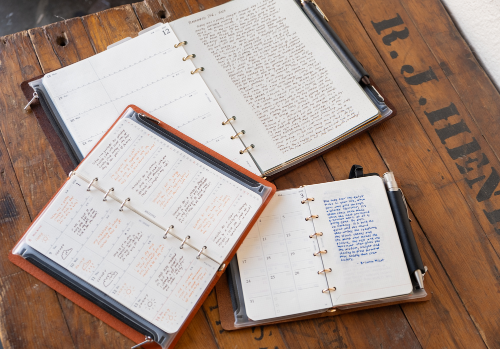 Three open journals with handwritten entries sit on a rustic wooden table. Notes are written in black, blue, and orange. Pens rest beside each journal.
