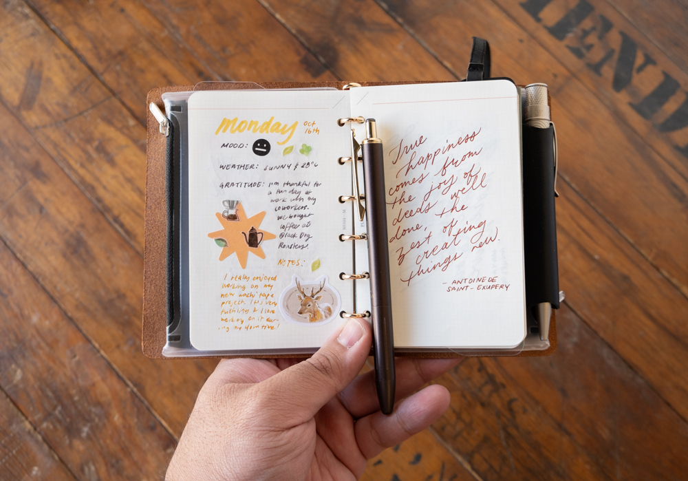 A hand holding an open leather-bound journal on a wooden table. The left page has colorful doodles and handwritten notes about mood and gratitude. The right page shows a motivational quote written in red ink. A pen is placed at the journal's center.