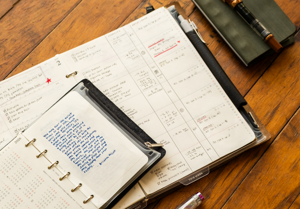 Open planner on a wooden table with handwritten notes, a black pen, and a smaller PLOTTER Leather Binder. The scene conveys organization and productivity.