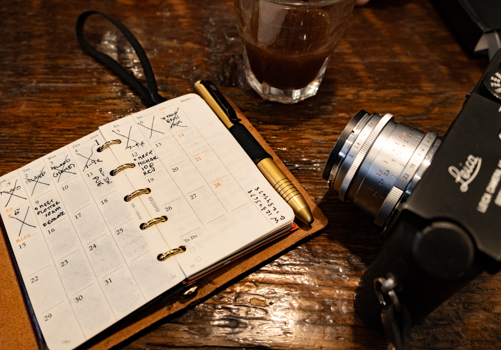 Open planner with crossed-out dates, a black and gold pen, a camera, and a glass of coffee on a rustic wooden table, conveying a busy, creative atmosphere.