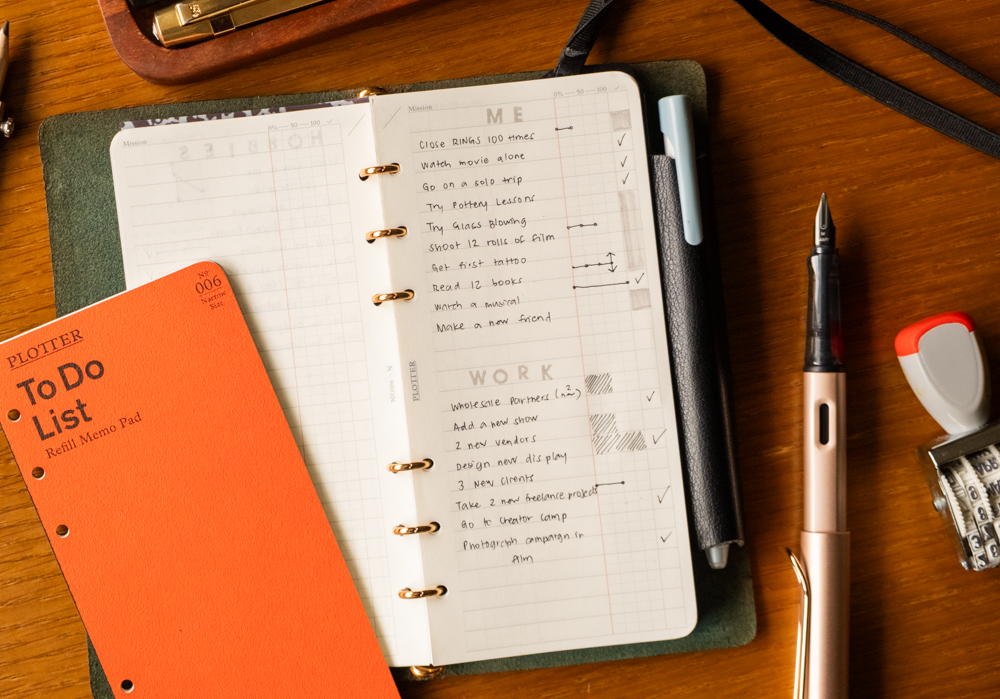 A planner and an orange to-do list pad on a wooden desk. The planner contains handwritten tasks under "Me" and "Work" with checkmarks. Nearby are pens and a date stamp. The scene conveys organization and productivity.