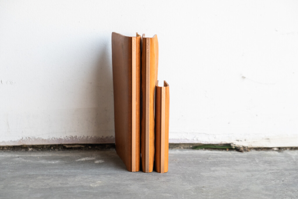 Three PLOTTER Leather Binders in Horween Predator Leather in Narrow Size, Bible Size and Mini 5 Size standing upright against a white wall and concrete floors.