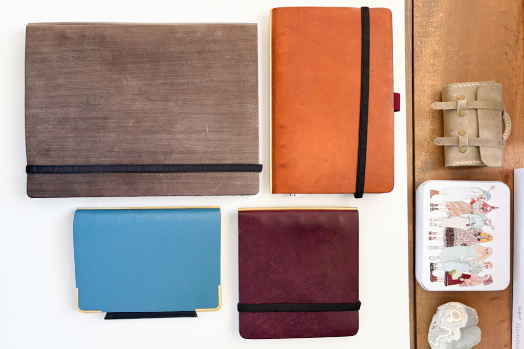 A neatly arranged collection of items on a wooden table includes PLOTTER Leather Binders in beige, brown, wine, and blue, a case of colorful markers, a small satchel, and decorative objects, conveying an organized and creative workspace.
