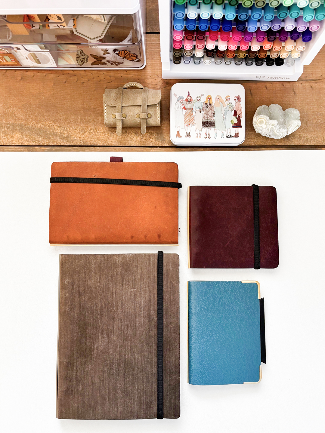A neatly arranged collection of items on a wooden table includes PLOTTER Leather Binders in beige, brown, wine, and blue, a case of colorful markers, a small satchel, and decorative objects, conveying an organized and creative workspace.