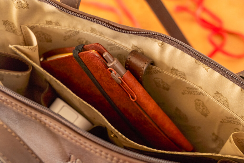 A red brown PLOTTER Leather Binder is inside a pocket in a leather shoulder bag.