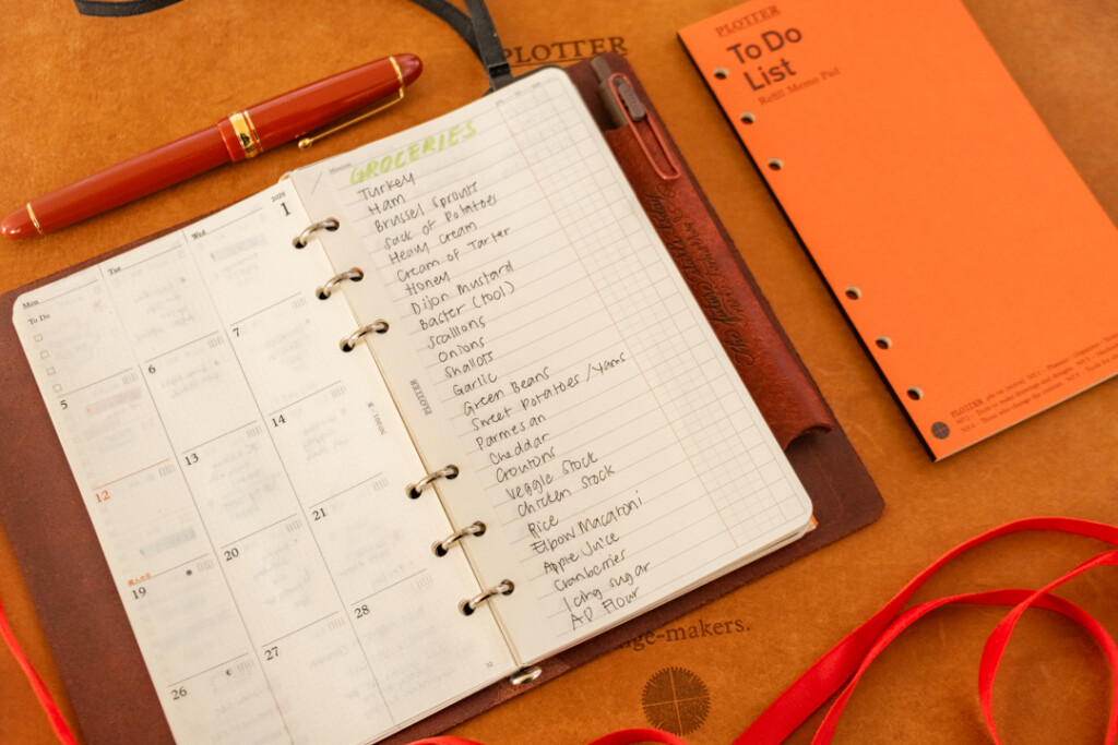 A PLOTTER Leather Binder open to a shopping list on a leather placemat with red ribbon and refill memo pads near the PLOTTER as decoration.