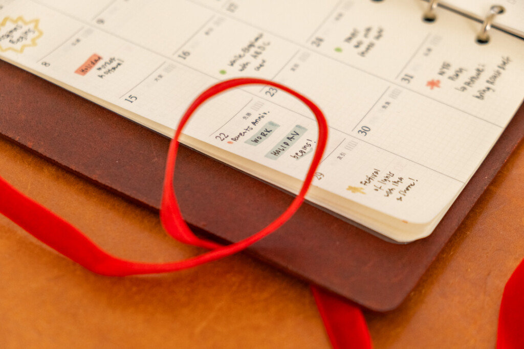 PLOTTER Leather Binder open to a monthly schedule with different holiday plans on a leather placemat with red ribbon nearby.
