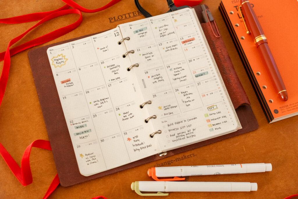 PLOTTER Leather Binder open to a monthly schedule with different holiday plans on a leather placemat with red ribbon and a mini Christmas tree near the PLOTTER as decoration.