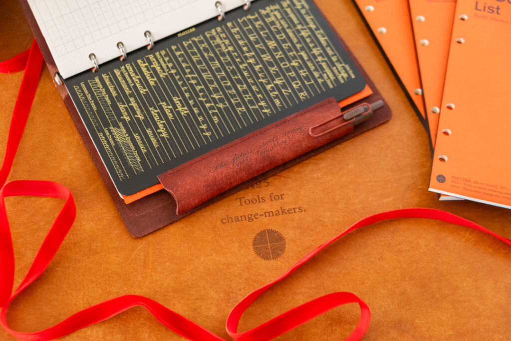 A Red Brown PLOTTER Leather Binder open to show the lifter with pen holder. The PLOTTER Leather Binder is on a leather placemat with red ribbon, and refill memo pads.