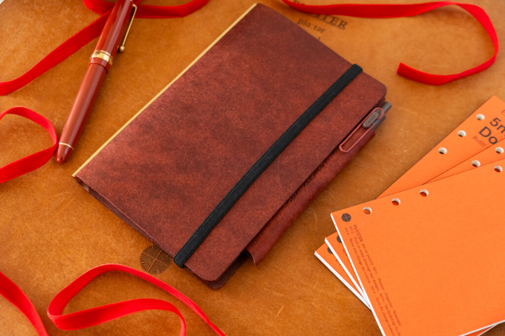 A Red Brown PLOTTER Leather Binder on a leather placemat with red ribbon, and refill memo pads.