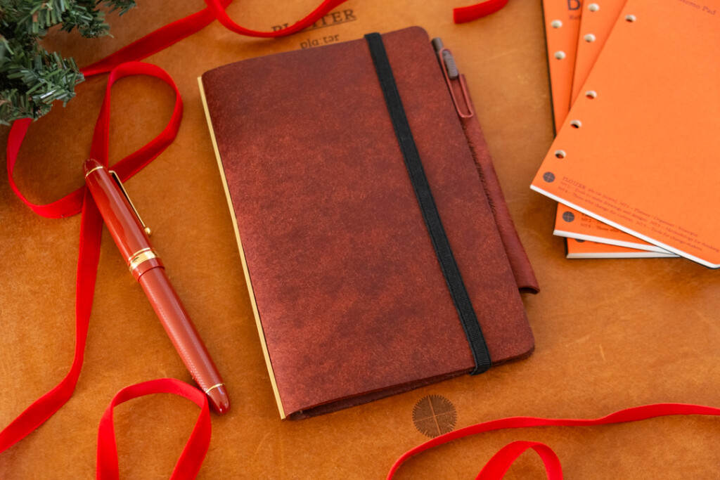A Red Brown PLOTTER Leather Binder on a leather placemat with red ribbon, refill memo pads, and a mini Christmas tree nearby.